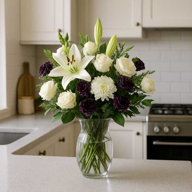 Vase-ready sympathy bouquet of white lily, cream roses, and purple carnations on a marble kitchen bench in a bright kitchen