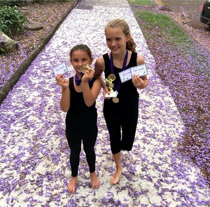 Asha and Ivy in Grafton holding their drama competition awards, standing on a footpath covered in fallen jacaranda flowers