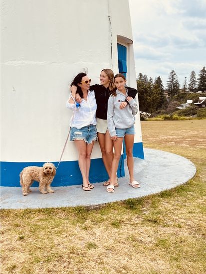 Siobhan with daughters Asha and Ivy and their dog Bindi standing at the base of the Clarence Head Lighthouse in Yamba, white tower with blue trim, Norfolk Pines visible in the background