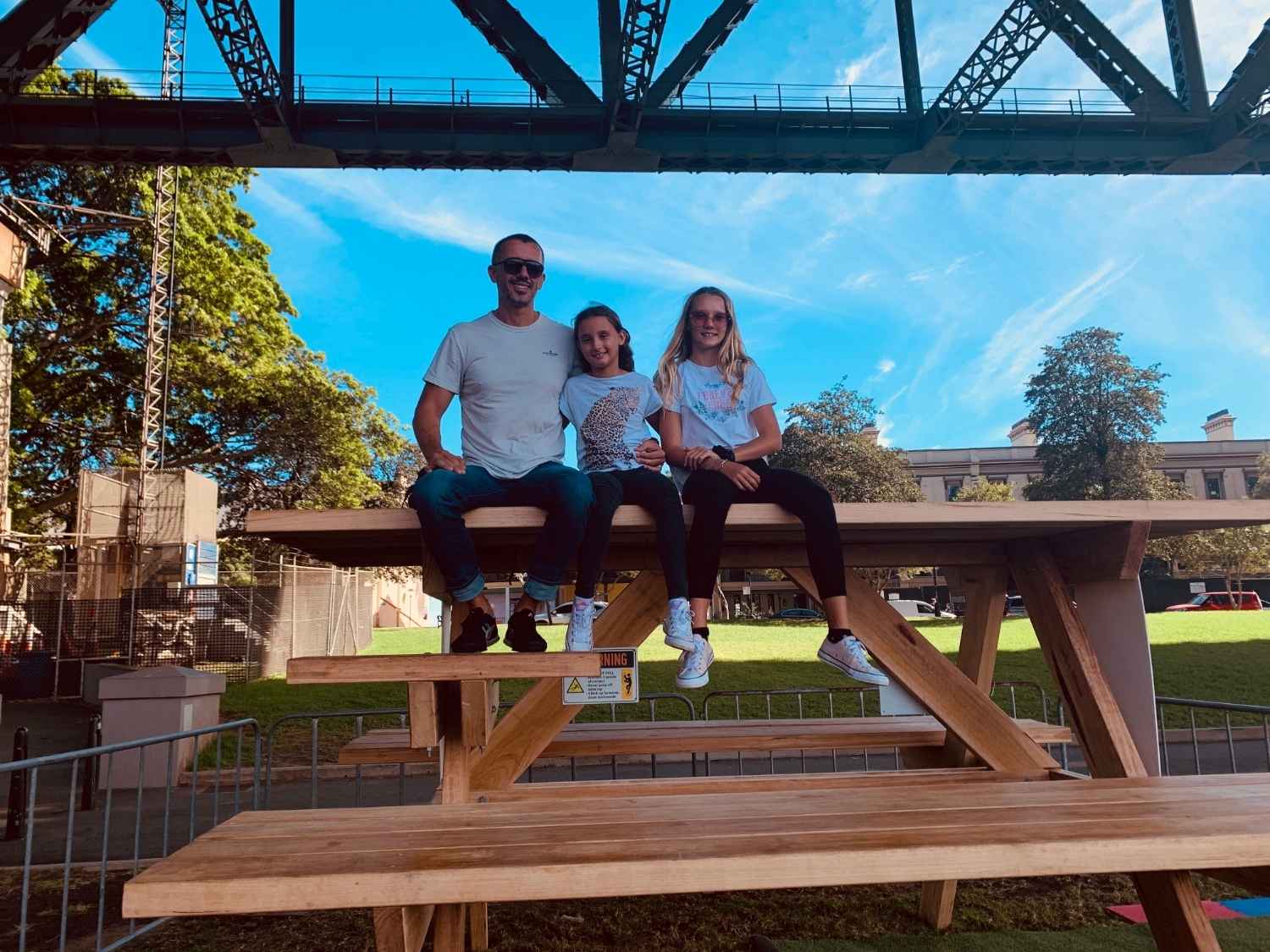 Andrew with daughters Asha and Ivy sitting on an oversized picnic table public artwork at The Rocks in Sydney, with the Harbour Bridge steel arches directly above them